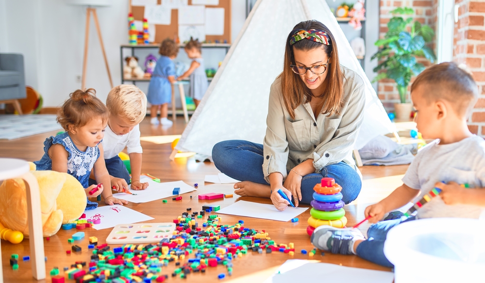 Beautiful teacher and group of toddlers playing around lots of toys at kindergarten